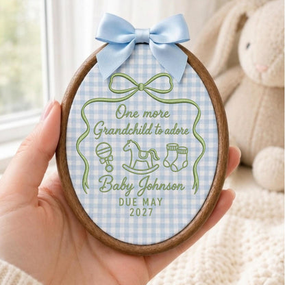 May include: An oval wooden embroidery hoop frame containing light blue and white gingham fabric. The fabric features green embroidered text reading One more Grandchild to adore, Baby Johnson, DUE MAY 2027, with icons of a rattle, rocking horse, and socks.
