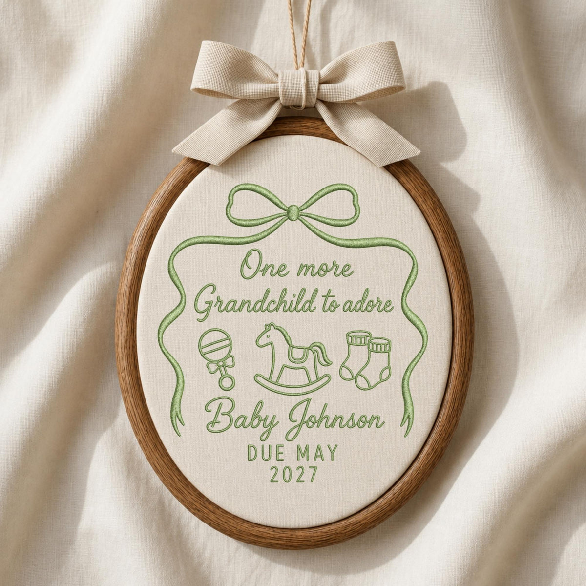 May include: An oval wooden embroidery hoop containing cream fabric with green stitched text reading One more Grandchild to adore, Baby Johnson, and DUE MAY 2027. The design includes a green stitched ribbon, rattle, rocking horse, and booties.