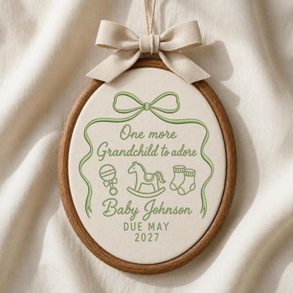 May include: An oval wooden embroidery hoop containing cream fabric with green stitched text reading One more Grandchild to adore, Baby Johnson, and DUE MAY 2027. The design includes a green stitched ribbon, rattle, rocking horse, and booties.