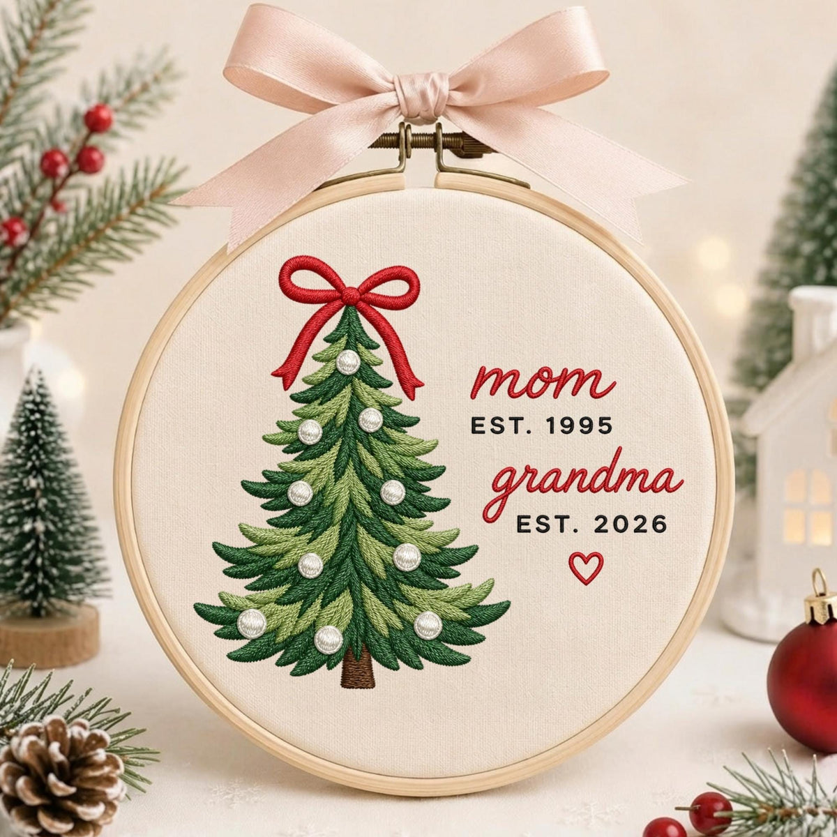 May include: A circular wooden embroidery hoop containing cream-colored fabric with a stitched green Christmas tree, a red bow, and white pearl accents. The text reads mom EST. 1995, grandma EST. 2026, with a small red heart. A light pink ribbon is tied at the top.