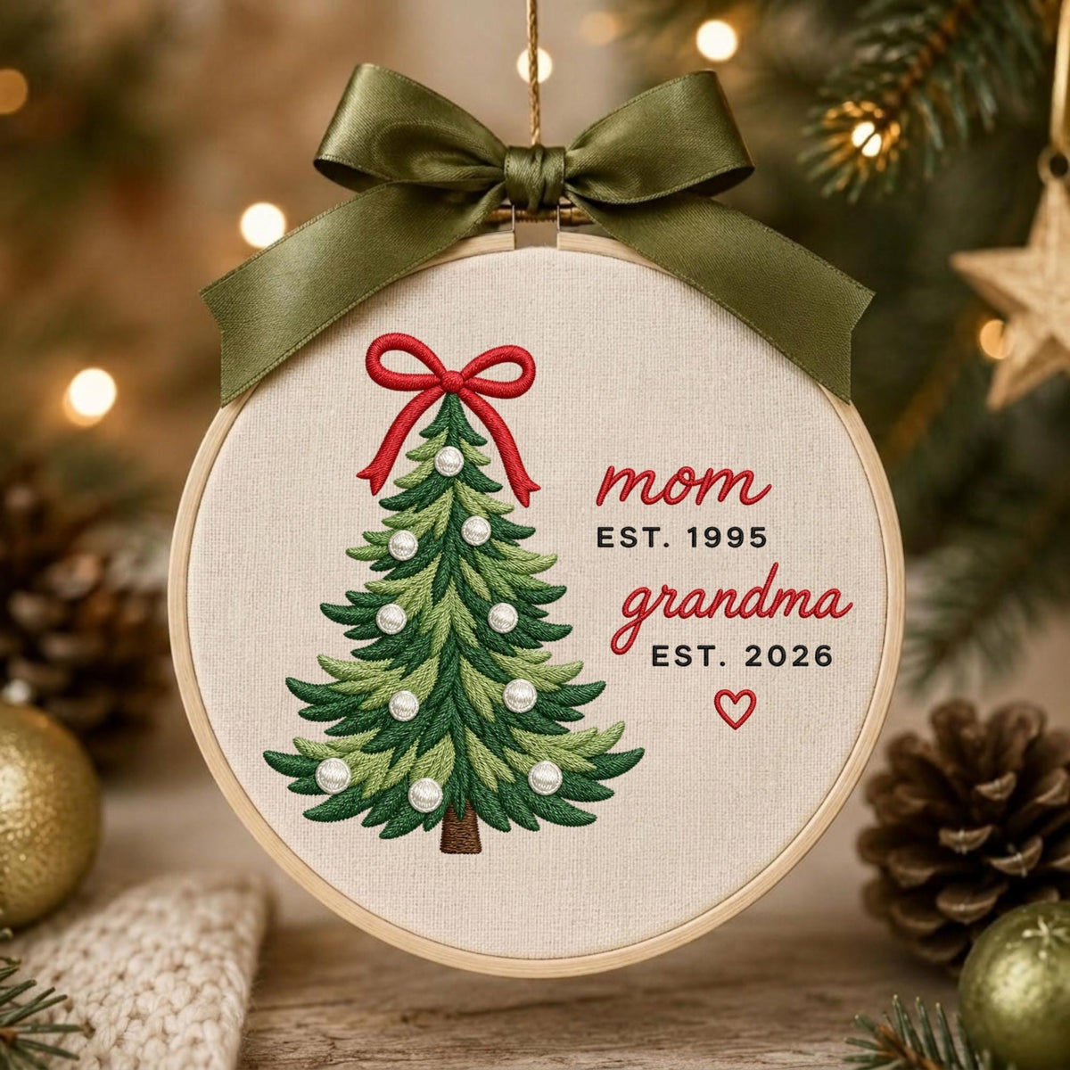 May include: A round wooden embroidery hoop ornament featuring a stitched green Christmas tree with white pearl accents and a red bow topper. The text reads mom EST. 1995, grandma EST. 2026, and a small red heart. An olive green satin ribbon is tied at the top.