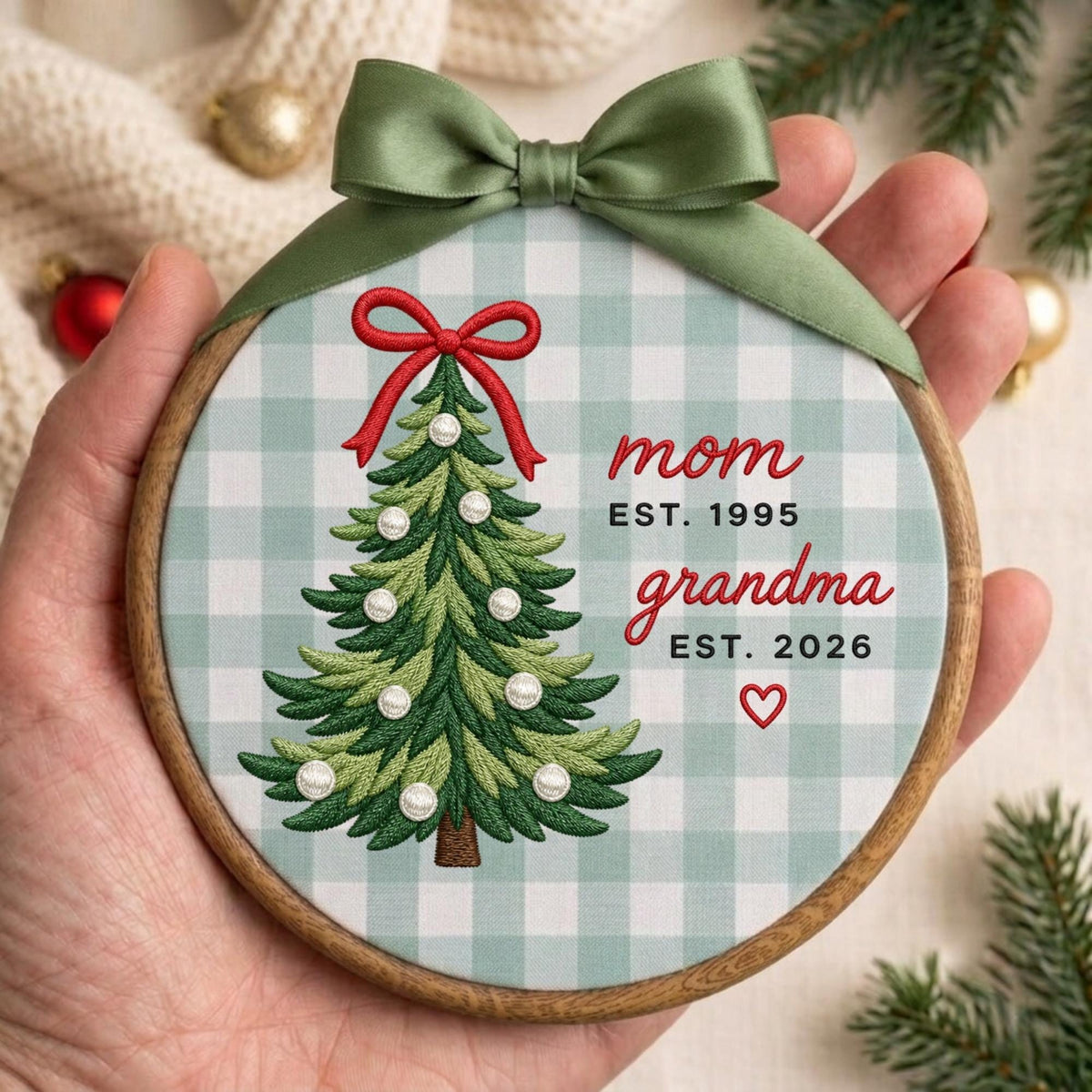 May include: A round wooden embroidery hoop ornament featuring a light green and white buffalo check fabric. An embroidered green Christmas tree with a red bow and white ornaments is centered. Text reads: mom EST. 1995, grandma EST. 2026, and a red heart.