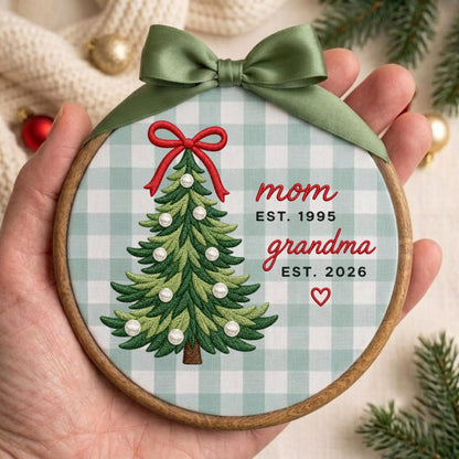May include: A round wooden embroidery hoop ornament featuring a light green and white buffalo check fabric. An embroidered green Christmas tree with a red bow and white ornaments is centered. Text reads: mom EST. 1995, grandma EST. 2026, and a red heart.
