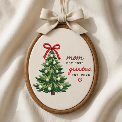 May include: An oval wooden embroidery hoop containing cream fabric with a stitched green Christmas tree decorated with white pearl-like beads and a red bow. The text reads mom EST. 1995, grandma EST. 2026, and a small red heart. A beige ribbon bow hangs from the top.