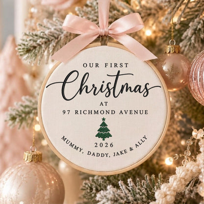 May include: A round wooden embroidery hoop ornament featuring cream fabric with black text reading OUR FIRST Christmas AT 97 RICHMOND AVENUE, a green embroidered pine tree, the year 2026, and the names MUMMY, DADDY, JAKE &amp; ALLY, topped with a pale pink satin ribbon bow.