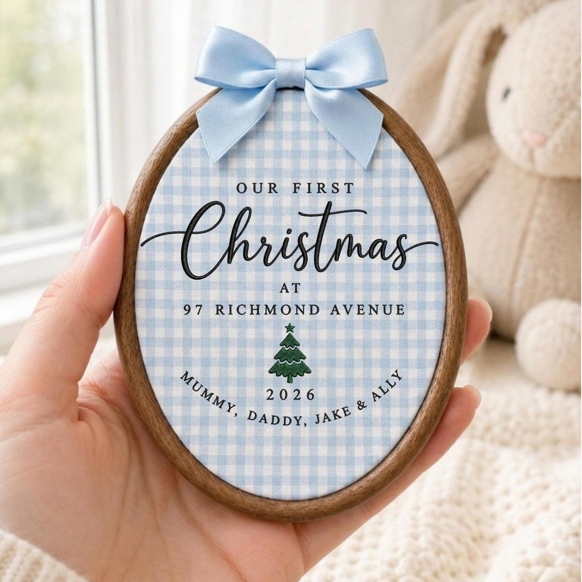 May include: An oval wooden embroidery hoop ornament featuring a light blue and white gingham fabric background. The text reads Our First Christmas at 97 Richmond Avenue, 2026, Mummy, Daddy, Jake &amp; Ally, with a green Christmas tree icon and a light blue satin bow.