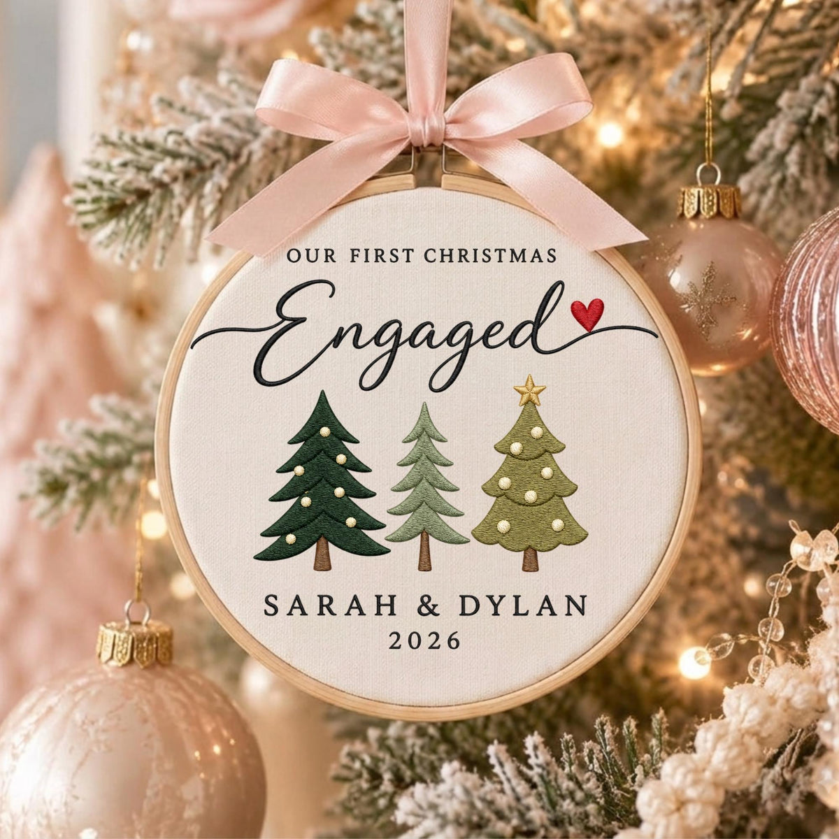May include: A round wooden embroidery hoop ornament featuring white fabric with the text OUR FIRST CHRISTMAS Engaged, a red heart, three embroidered pine trees, and the names SARAH &amp; DYLAN with the year 2026, topped with a light pink satin bow.