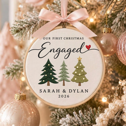 May include: A round wooden embroidery hoop ornament featuring white fabric with the text OUR FIRST CHRISTMAS Engaged, a red heart, three embroidered pine trees, and the names SARAH &amp; DYLAN with the year 2026, topped with a light pink satin bow.