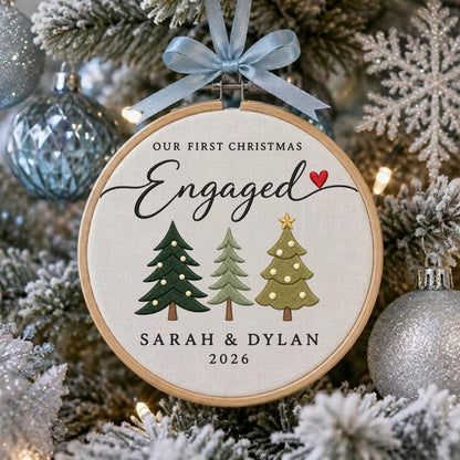 May include: A round wooden embroidery hoop ornament featuring the embroidered text OUR FIRST CHRISTMAS Engaged with a red heart, three stylized Christmas trees in shades of green, and the names SARAH &amp; DYLAN with the year 2026, topped with a light blue ribbon.