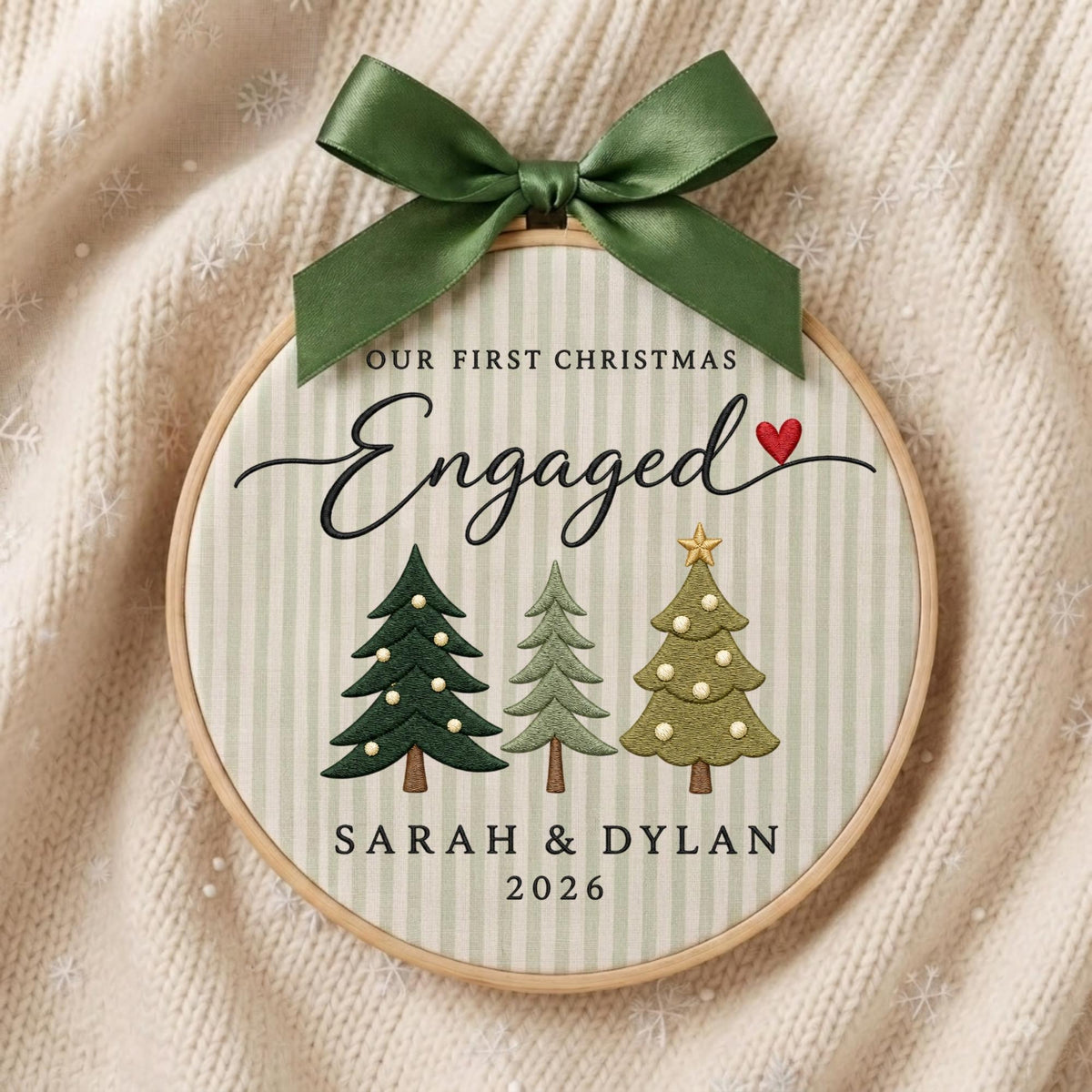 May include: A circular wooden embroidery hoop ornament featuring a light green and white striped fabric background. It displays the text "OUR FIRST CHRISTMAS Engaged" with a small red heart, three stylized Christmas trees, and "SARAH &amp; DYLAN 2026". A dark green satin ribbon bow is tied at the top.