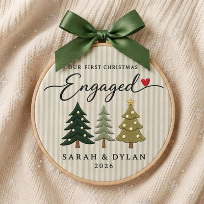May include: A circular wooden embroidery hoop ornament featuring a light green and white striped fabric background. It displays the text "OUR FIRST CHRISTMAS Engaged" with a small red heart, three stylized Christmas trees, and "SARAH &amp; DYLAN 2026". A dark green satin ribbon bow is tied at the top.