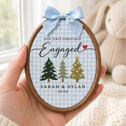 May include: An oval wooden embroidery hoop ornament featuring a light blue and white gingham fabric background. It displays the text OUR FIRST CHRISTMAS Engaged with a red heart, three illustrated Christmas trees, and the names SARAH &amp; DYLAN 2026.