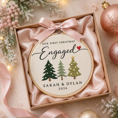May include: A round wooden embroidery hoop ornament featuring the text "OUR FIRST CHRISTMAS Engaged" with a small red heart, three embroidered Christmas trees, and the names "SARAH &amp; DYLAN 2026" in a gift box with a light pink satin ribbon bow.