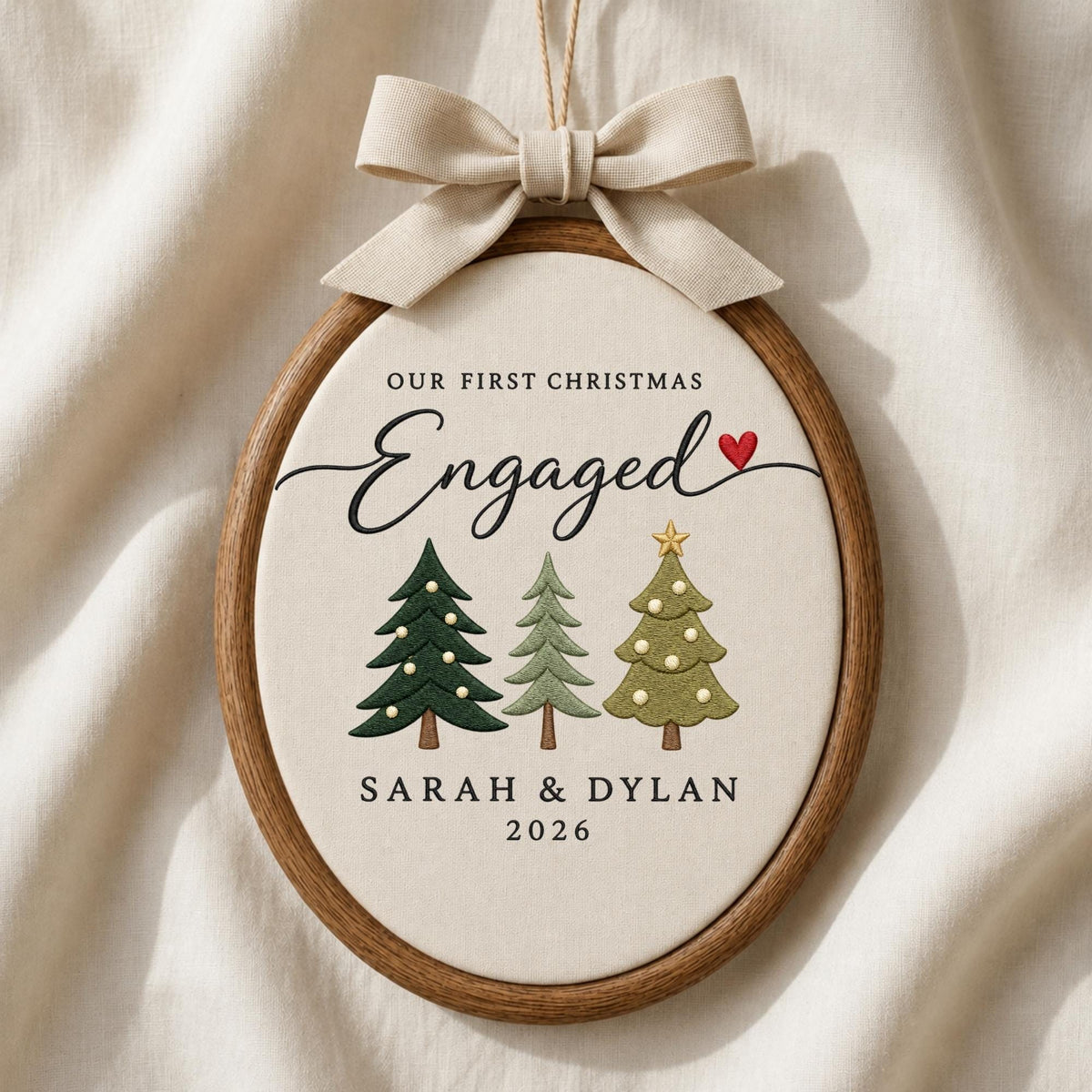 May include: An oval wooden embroidery hoop ornament featuring cream fabric with the text OUR FIRST CHRISTMAS Engaged, a red heart, and three embroidered Christmas trees. The names SARAH &amp; DYLAN and the year 2026 are printed below, topped with a beige bow.