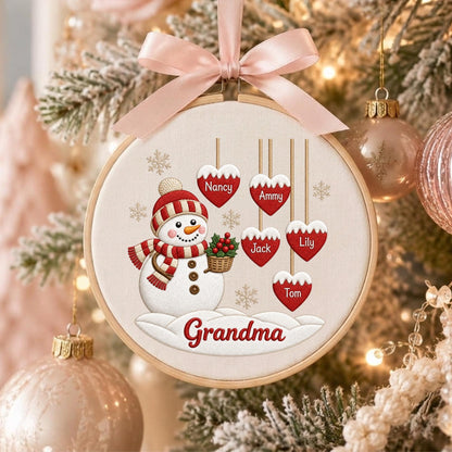 May include: A round embroidery hoop ornament featuring a snowman wearing a red and white striped hat and scarf, holding a basket of holly. Five red heart-shaped ornaments hang above with the names Nancy, Ammy, Jack, Lily, and Tom. Grandma is written below.