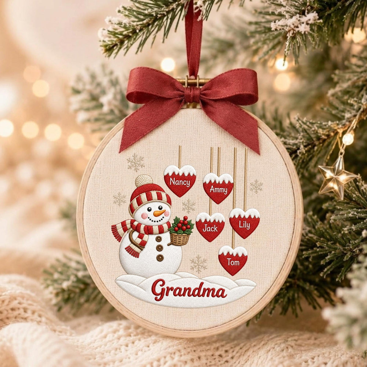 May include: A round wooden embroidery hoop ornament featuring a snowman wearing a red and white striped hat and scarf, holding a basket of holly. Five red heart-shaped tags hang above, labeled Nancy, Ammy, Jack, Lily, and Tom. Grandma is written below.