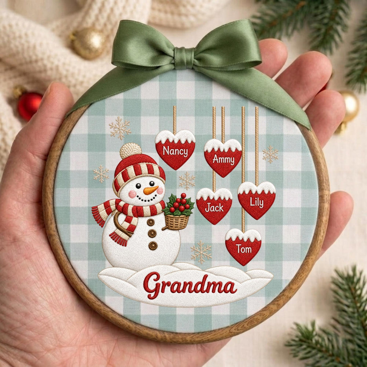 May include: A round wooden embroidery hoop ornament featuring a light blue and white gingham fabric background. A snowman with a red and white striped hat and scarf holds a basket of holly. Five red heart ornaments hang above with the names Nancy, Ammy, Jack, Lily, and Tom. The word Grandma is printed in red on a snowdrift at the bottom, topped with a sage green satin ribbon bow.