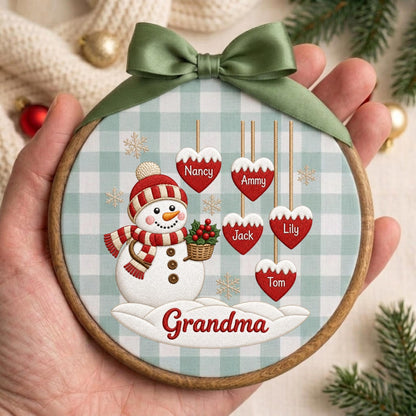 May include: A round wooden embroidery hoop ornament featuring a light blue and white gingham fabric background. A snowman with a red and white striped hat and scarf holds a basket of holly. Five red heart ornaments hang above with the names Nancy, Ammy, Jack, Lily, and Tom. The word Grandma is printed in red on a snowdrift at the bottom, topped with a sage green satin ribbon bow.