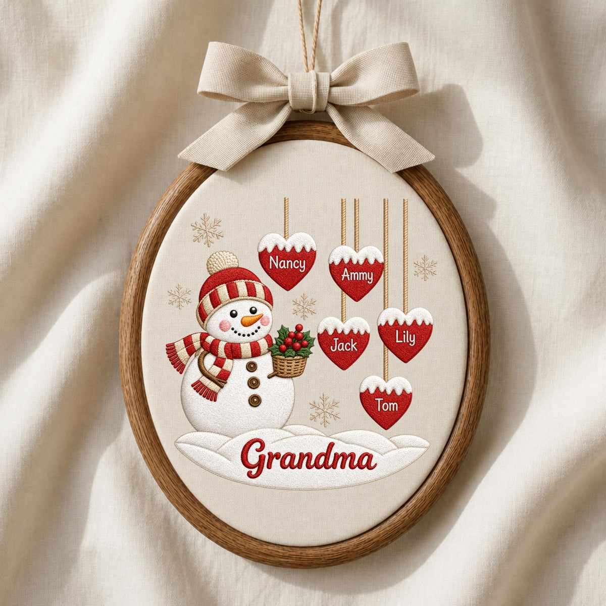 May include: An oval wooden embroidery hoop frame containing a fabric panel with a snowman illustration wearing a red and white striped hat and scarf. Five red heart ornaments hang above, labeled Nancy, Ammy, Jack, Lily, and Tom. Grandma is written below.