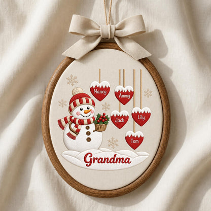 May include: An oval wooden embroidery hoop frame containing a fabric panel with a snowman illustration wearing a red and white striped hat and scarf. Five red heart ornaments hang above, labeled Nancy, Ammy, Jack, Lily, and Tom. Grandma is written below.