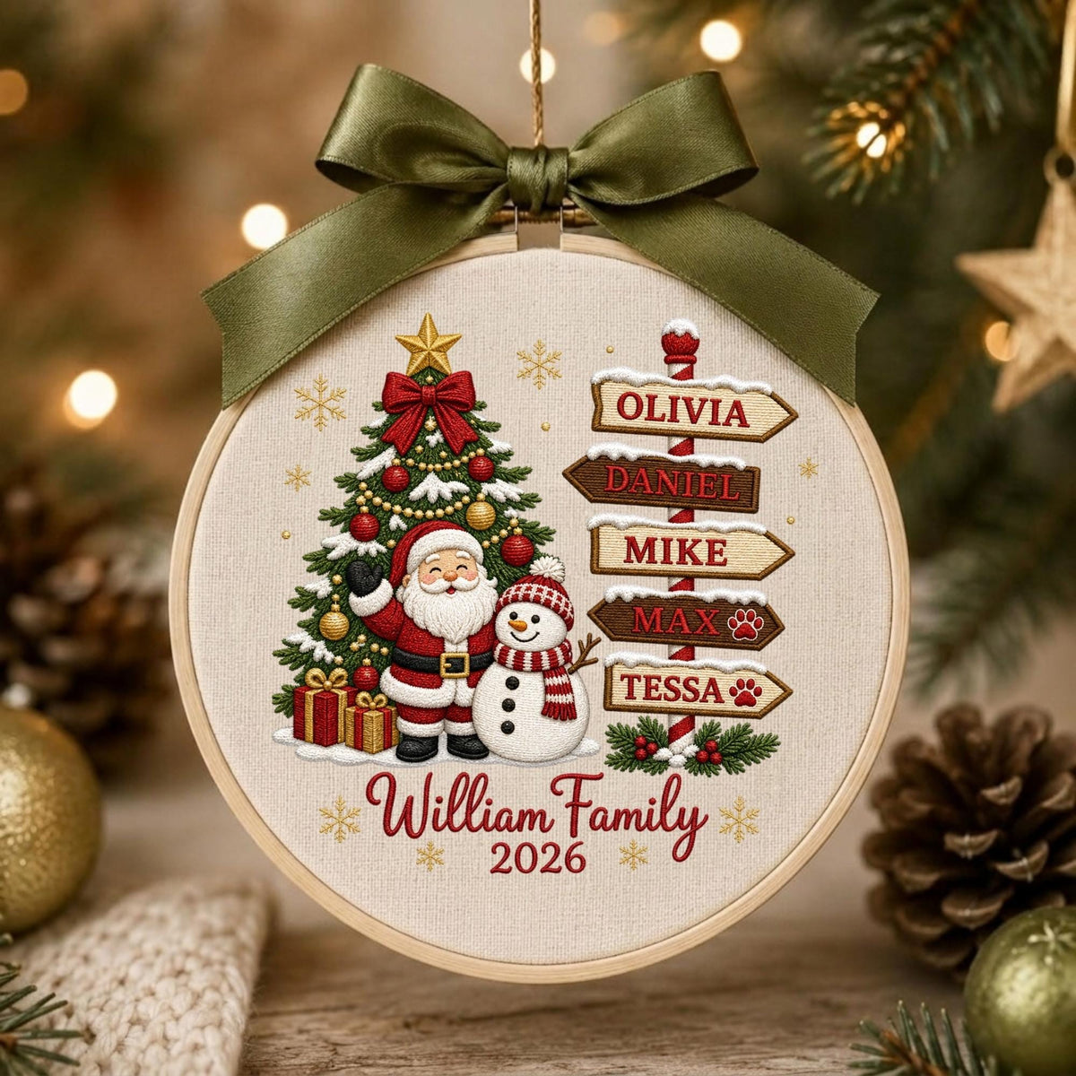 May include: A round embroidery hoop ornament featuring a cream fabric background with a printed design of a Christmas tree, Santa Claus, a snowman, and a signpost with the names Olivia, Daniel, Mike, Max, and Tessa. It includes a dark green ribbon bow.