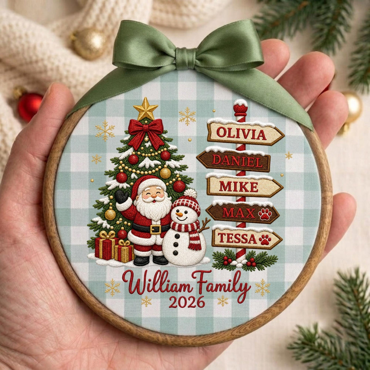 May include: A round wooden embroidery hoop ornament featuring a light blue and white buffalo check background. The design includes a decorated Christmas tree, Santa Claus, a snowman, and a signpost with the names Olivia, Daniel, Mike, Max, and Tessa. The text reads William Family 2026, topped with a sage green satin ribbon bow.