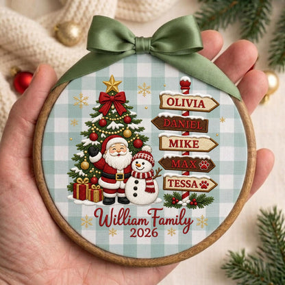May include: A round wooden embroidery hoop ornament featuring a light blue and white buffalo check background. The design includes a decorated Christmas tree, Santa Claus, a snowman, and a signpost with the names Olivia, Daniel, Mike, Max, and Tessa. The text reads William Family 2026, topped with a sage green satin ribbon bow.