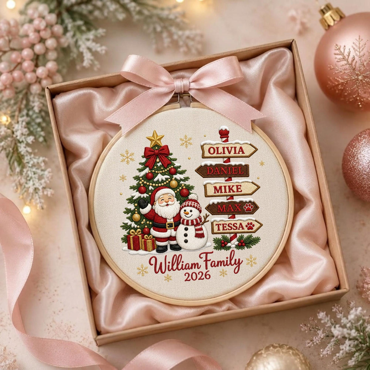 May include: A round embroidery hoop featuring a Christmas tree, Santa Claus, and a snowman. A wooden signpost displays the names Olivia, Daniel, Mike, Max, and Tessa. The text William Family 2026 is printed below. A light pink satin ribbon is tied at the top.