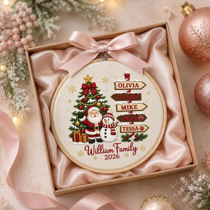 May include: A round embroidery hoop featuring a Christmas tree, Santa Claus, and a snowman. A wooden signpost displays the names Olivia, Daniel, Mike, Max, and Tessa. The text William Family 2026 is printed below. A light pink satin ribbon is tied at the top.