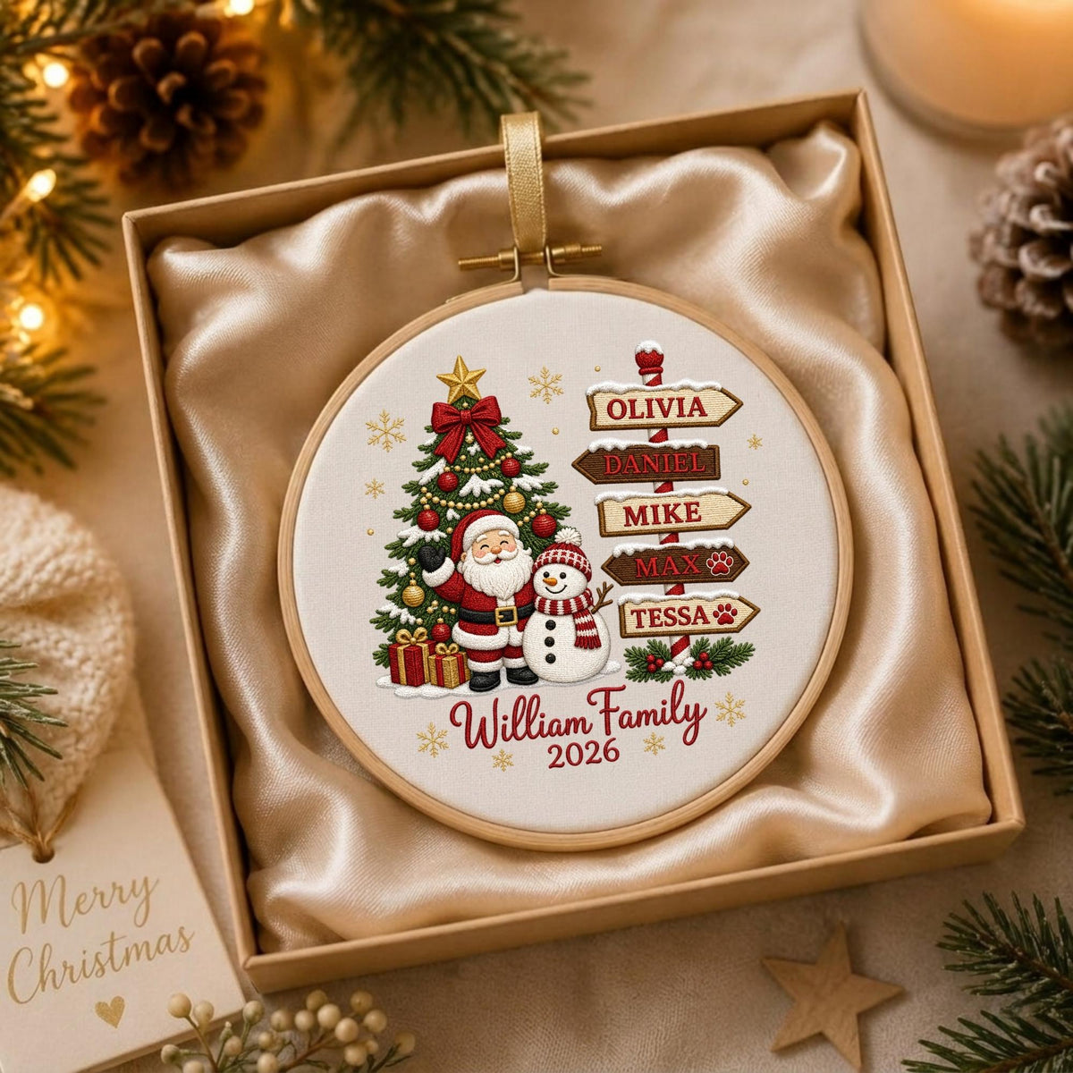May include: A round Christmas ornament in a wooden embroidery hoop featuring a printed design of Santa Claus, a snowman, a decorated tree, and a signpost with the names Olivia, Daniel, Mike, Max, and Tessa, plus the text William Family 2026.
