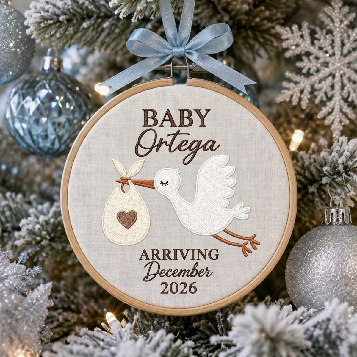 May include: A round wooden embroidery hoop ornament featuring a white fabric center with a brown embroidered stork carrying a bundle with a heart. The text reads BABY Ortega ARRIVING December 2026. A light blue ribbon bow is tied at the top.