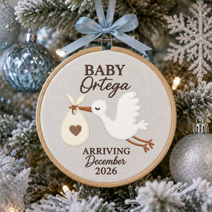 May include: A round wooden embroidery hoop ornament featuring a white fabric center with a brown embroidered stork carrying a bundle with a heart. The text reads BABY Ortega ARRIVING December 2026. A light blue ribbon bow is tied at the top.