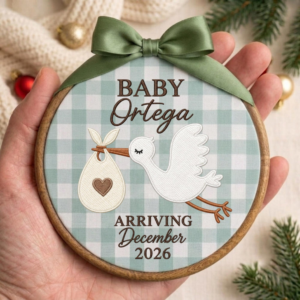 May include: A round wooden embroidery hoop ornament featuring a light green and white gingham fabric background. It displays a white stork carrying a bundle with a brown heart, and the text BABY Ortega ARRIVING December 2026. A sage green satin ribbon bow is attached to the top.