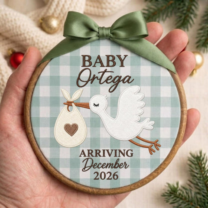 May include: A round wooden embroidery hoop ornament featuring a light green and white gingham fabric background. It displays a white stork carrying a bundle with a brown heart, and the text BABY Ortega ARRIVING December 2026. A sage green satin ribbon bow is attached to the top.