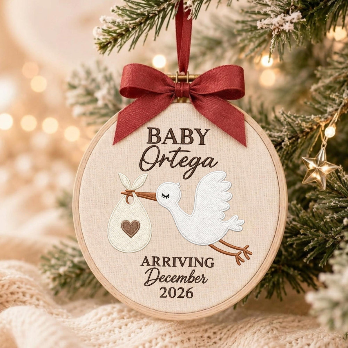 May include: A round wooden embroidery hoop ornament featuring a white stork carrying a bundle with a brown heart. The text reads BABY Ortega ARRIVING December 2026. A dark red satin ribbon bow is tied at the top for hanging on a Christmas tree.
