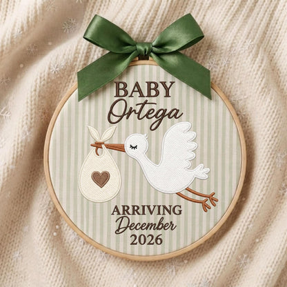 May include: A circular wooden embroidery hoop containing fabric with vertical light green stripes. It features an embroidered white stork carrying a bundle with a brown heart, the text BABY Ortega, and the text ARRIVING December 2026, topped with a dark green satin bow.