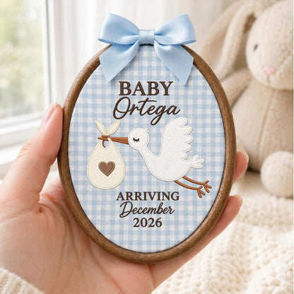 May include: An oval wooden embroidery hoop frame containing fabric with a light blue gingham pattern. It features an embroidered white stork carrying a bundle with a brown heart, and text reading BABY Ortega ARRIVING December 2026, topped with a light blue satin bow.