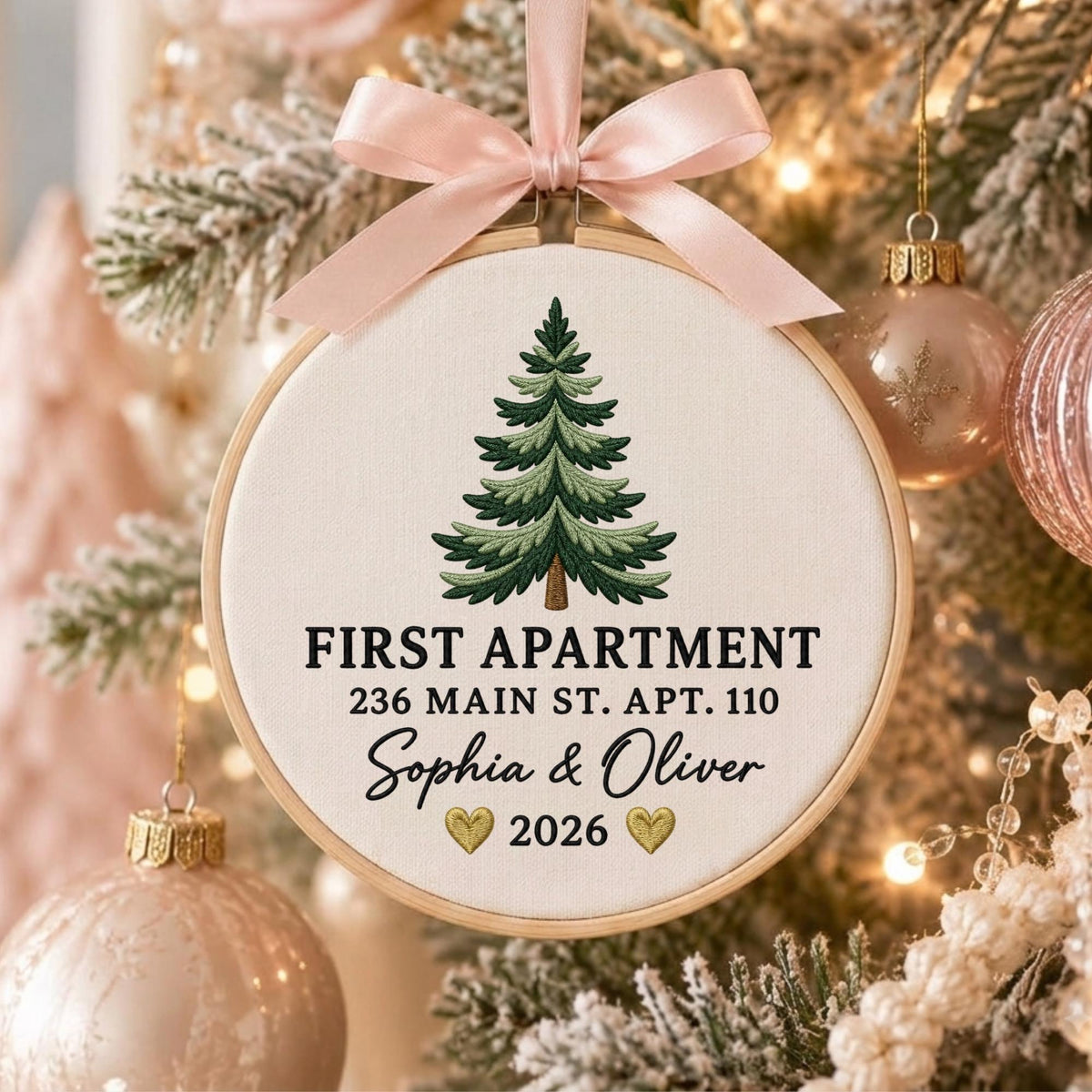 May include: A round wooden embroidery hoop ornament featuring a light-colored fabric center with an embroidered green pine tree. The text reads FIRST APARTMENT, 236 MAIN ST. APT. 110, Sophia &amp; Oliver, and 2026 with two gold hearts. A pink satin ribbon bow.
