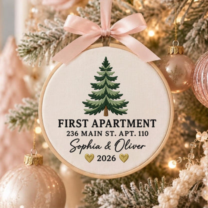 May include: A round wooden embroidery hoop ornament featuring a light-colored fabric center with an embroidered green pine tree. The text reads FIRST APARTMENT, 236 MAIN ST. APT. 110, Sophia &amp; Oliver, and 2026 with two gold hearts. A pink satin ribbon bow.