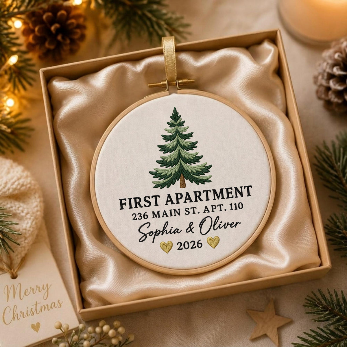 May include: A round embroidery hoop ornament featuring a stitched green pine tree, the text FIRST APARTMENT 236 MAIN ST. APT. 110, the names Sophia &amp; Oliver, two small gold hearts, and the year 2026, presented in a gift box with satin lining.
