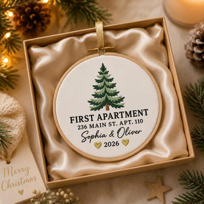 May include: A round embroidery hoop ornament featuring a stitched green pine tree, the text FIRST APARTMENT 236 MAIN ST. APT. 110, the names Sophia &amp; Oliver, two small gold hearts, and the year 2026, presented in a gift box with satin lining.