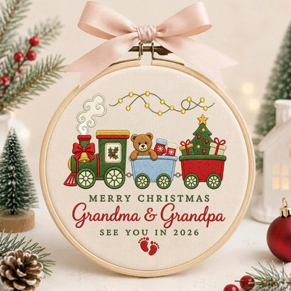 May include: A circular wooden embroidery hoop containing fabric with a stitched Christmas train design. The train features a green engine, a blue car with a teddy bear, and a red car with a tree. Text reads: MERRY CHRISTMAS Grandma &amp; Grandpa SEE YOU IN 2026, with red baby footprints below and a pink ribbon at the top.