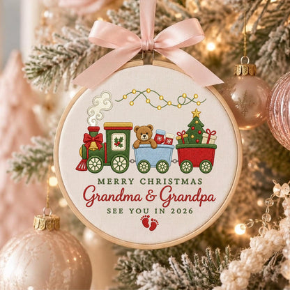 May include: A round embroidery hoop ornament featuring a train graphic with a teddy bear, a Christmas tree, and gifts. The text reads MERRY CHRISTMAS Grandma &amp; Grandpa SEE YOU IN 2026 with red baby footprints. A light pink satin ribbon bow is at the top.