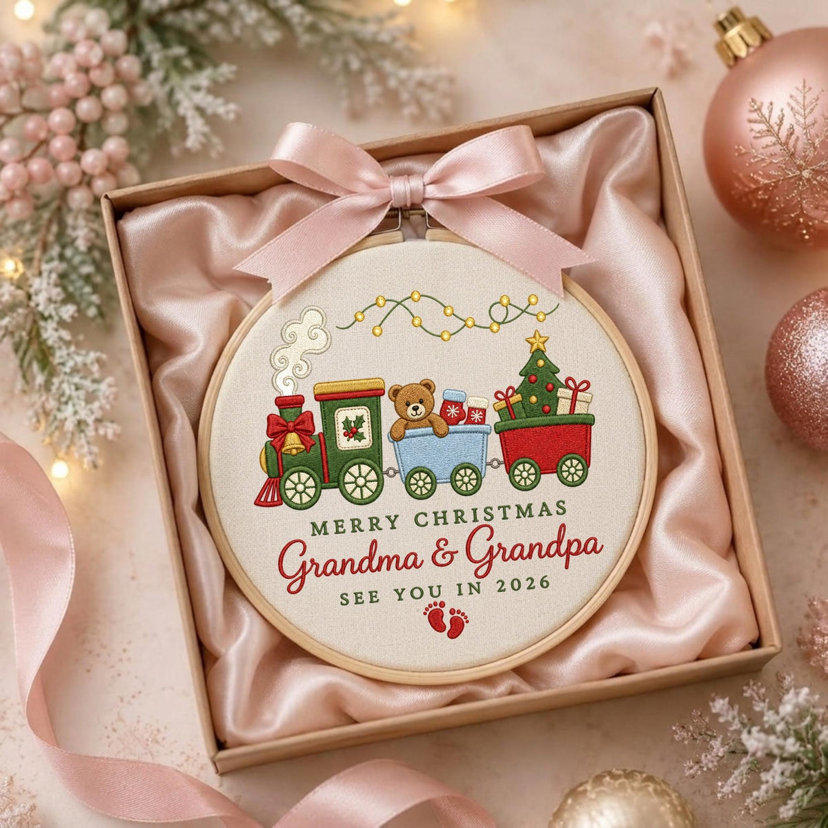 May include: A circular embroidery hoop featuring a stitched Christmas train with a teddy bear, gifts, and a tree. The text reads MERRY CHRISTMAS Grandma &amp; Grandpa SEE YOU IN 2026 with small red footprints. The hoop is topped with a light pink satin bow.