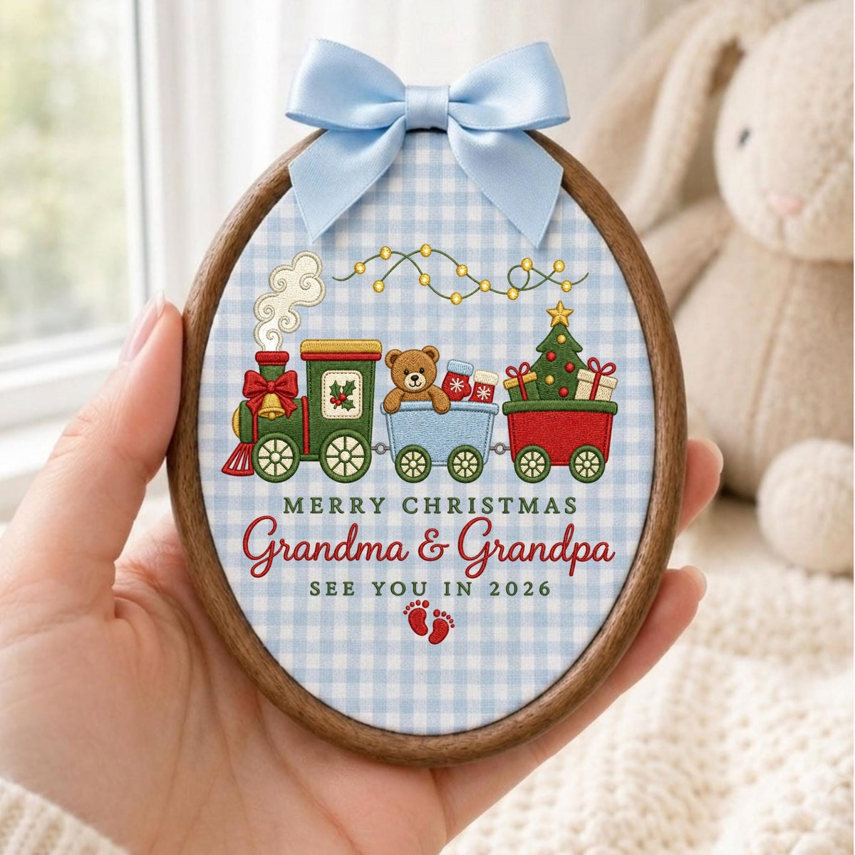 May include: An oval wooden embroidery hoop frame containing fabric with a light blue gingham pattern. The design features a colorful toy train, a teddy bear, a Christmas tree, and the text Merry Christmas Grandma &amp; Grandpa See You In 2026.