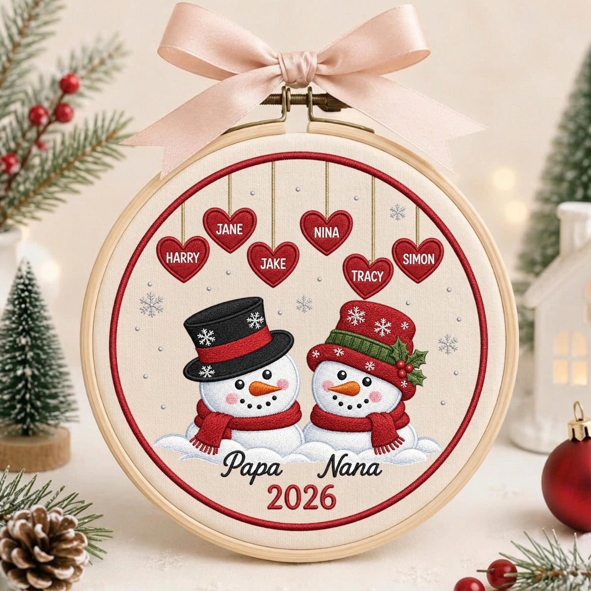May include: A circular embroidery hoop featuring a cream fabric background with two snowmen wearing hats and scarves. Above them, six red hearts hang with the names Harry, Jane, Jake, Nina, Tracy, and Simon. Below, the text reads Papa Nana 2026.