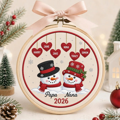 May include: A circular embroidery hoop featuring a cream fabric background with two snowmen wearing hats and scarves. Above them, six red hearts hang with the names Harry, Jane, Jake, Nina, Tracy, and Simon. Below, the text reads Papa Nana 2026.