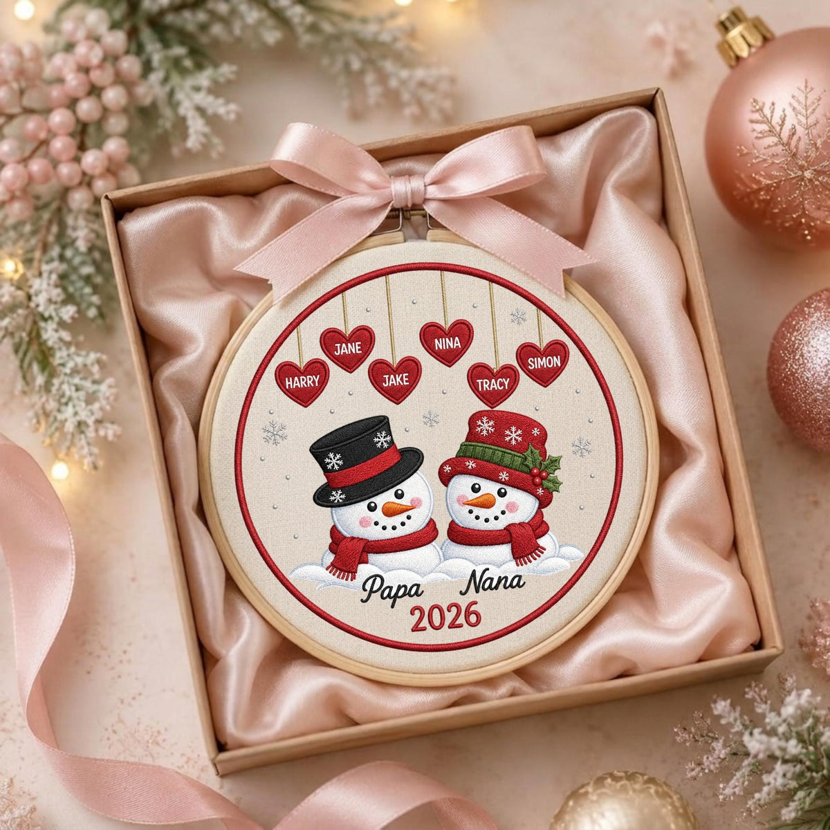 May include: A round embroidery hoop featuring two snowmen wearing hats and scarves, with six hanging red hearts labeled Harry, Jane, Jake, Nina, Tracy, and Simon. The text Papa, Nana, and 2026 is embroidered below. A pink satin bow sits on top.