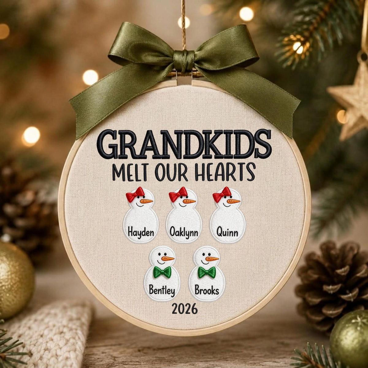 May include: A round embroidery hoop ornament featuring the embroidered text GRANDKIDS MELT OUR HEARTS and the year 2026. It displays five snowman figures with names Hayden, Oaklynn, Quinn, Bentley, and Brooks, topped with an olive green satin bow.