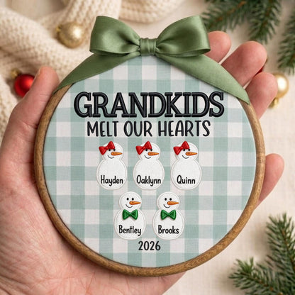 May include: A round wooden embroidery hoop ornament featuring a light green and white buffalo plaid fabric background. It displays the embroidered text GRANDKIDS MELT OUR HEARTS, five snowman figures with names, and the year 2026, topped with a green ribbon.